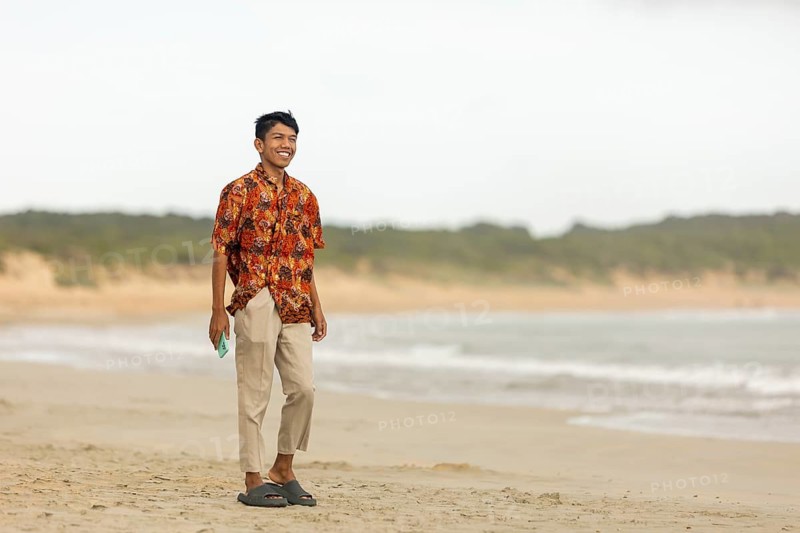Smiling Indonesian man wearing vibrant batik shirt, walking along an Australian beach. Sandy shore, ocean waves, and distant greenery create a serene backdrop