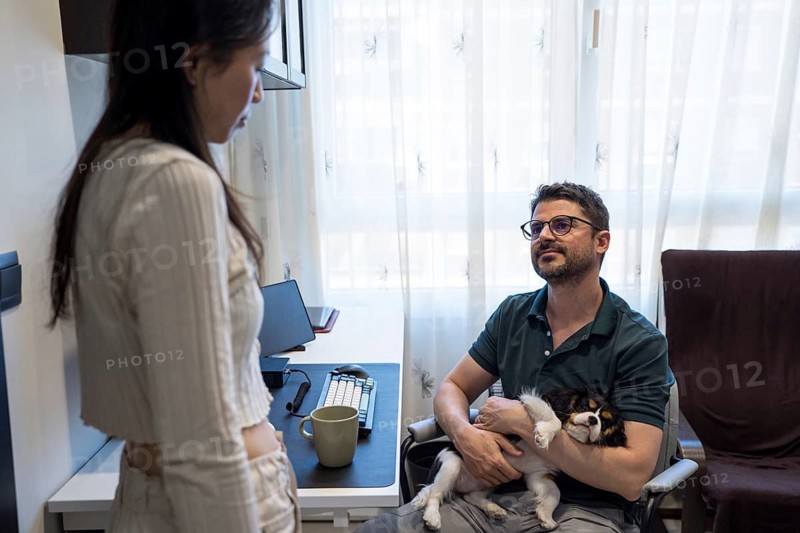 A diverse couple shares a warm moment at home, one seated with a small dog in an office chair, while the other stands nearby, creating a cozy, relaxed atmosphere