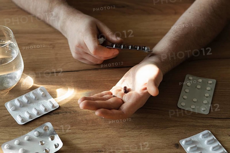 A man holds pills over a wooden table, with pill blister packs and a glass of water nearby, illustrating daily medication routine and healthcare at home