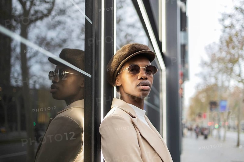 A stylish black woman in urban attire leans against a reflective glass wall, embodying confidence and modern fashion in a bustling cityscape
