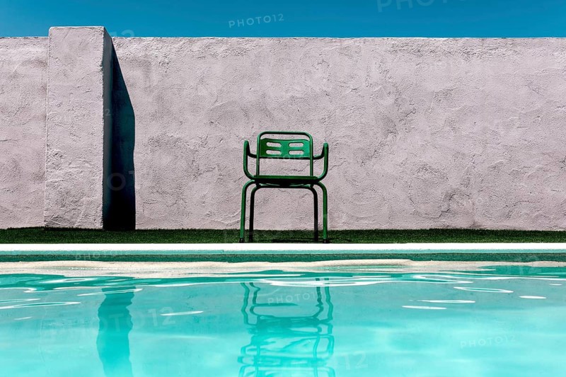 A vibrant green chair sits beside a serene pool, contrasting a textured concrete wall under a clear blue summer sky, creating a refreshing, aesthetic outdoor decor