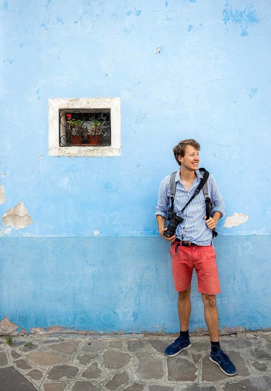 Young man in front of blue house wall with camera, small window with red flower, Burano Island, Venice, Veneto, Italy