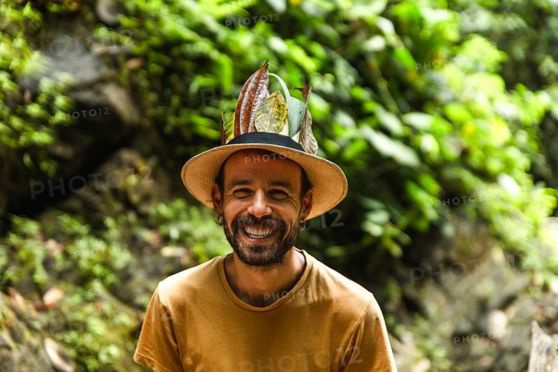 A cheerful Spanish young man beams joyfully in the Choco Andino rainforest of Ecuador, adorned with a unique hat decorated with tropical leaves, highlighting a true connection with the lush, biodiverse environment