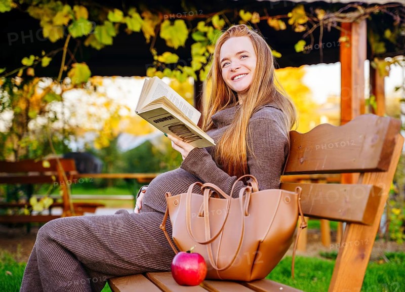 A serene pregnant woman with beautiful red hair sits on a wooden bench in an autumn park, engrossed in a book. The colorful foliage and warm sunlight create a peaceful setting
