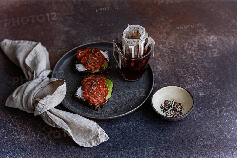 A rustic breakfast scene featuring whole wheat bread topped with pepperoni slices and avocado, accompanied by a steaming cup of drip coffee in a stylish setting