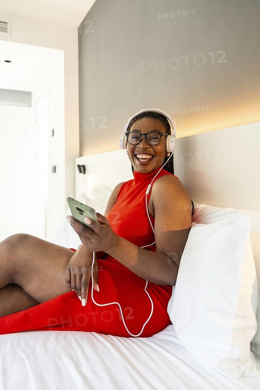Happy businesswoman wearing headphones, listening to music on smartphone while relaxing on hotel bed during business trip