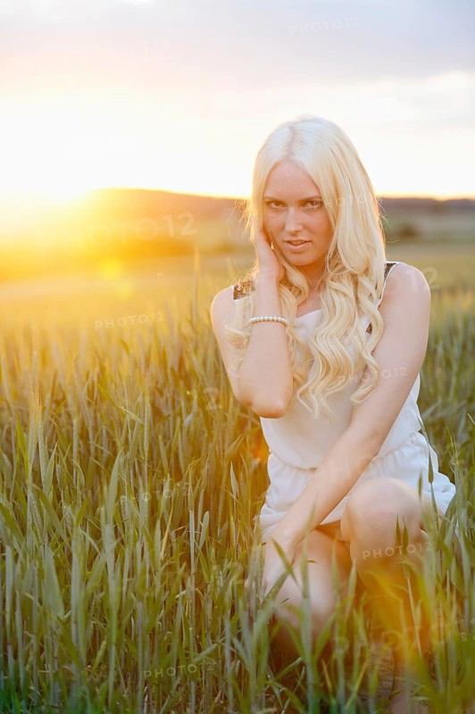 A blonde woman kneels in a grassy field at sunset, illuminated by warm sunlight