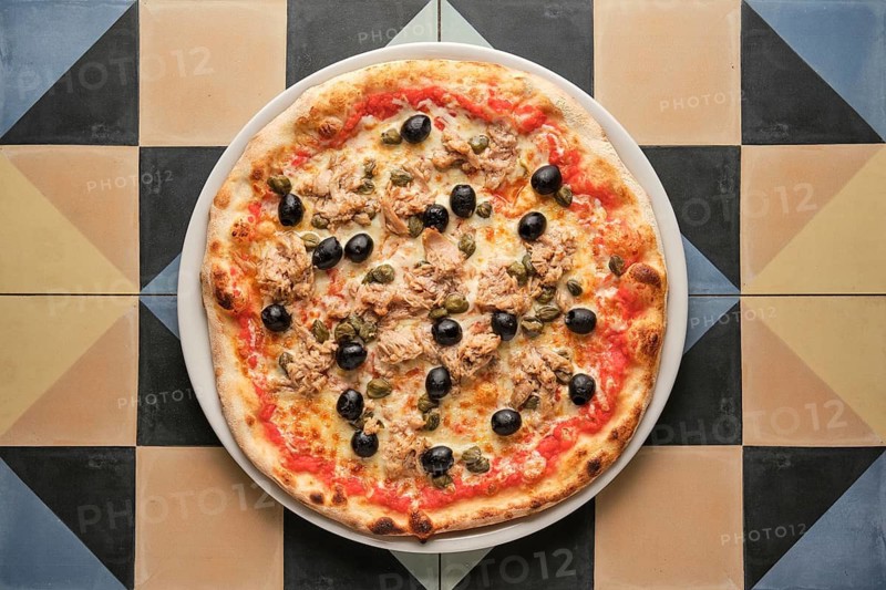 Overhead shot of a tuna pizza with capers and black olives, served on a white plate over colorful square tiles