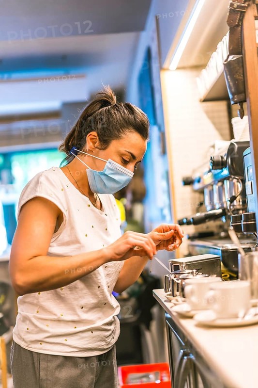 Photo session with a waitress with a face mask in a bar. New normality, security measures after the coronavirus pandemic, social distance, covid-19. Preparing a cut of coffee with milk with the machin