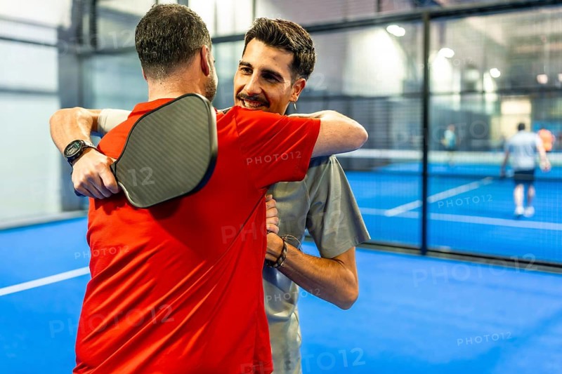 Two male friends embracing each other on a blue indoor court, smiling and showing good sportsmanship during a pickleball match, celebrating their camaraderie and athletic connection