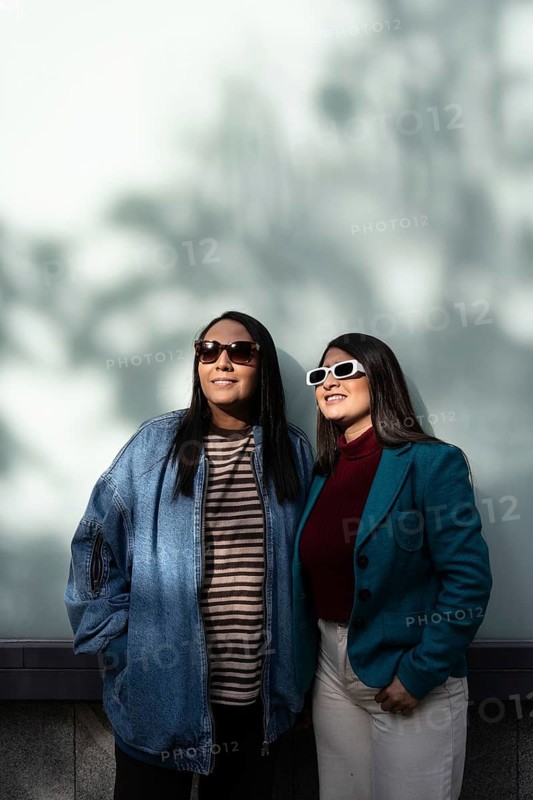 A Latin lesbian couple stands close, basking in the sun. One wears a denim jacket, the other a teal blazer, both sporting sunglasses. The backdrop is a soft, shadowed wall