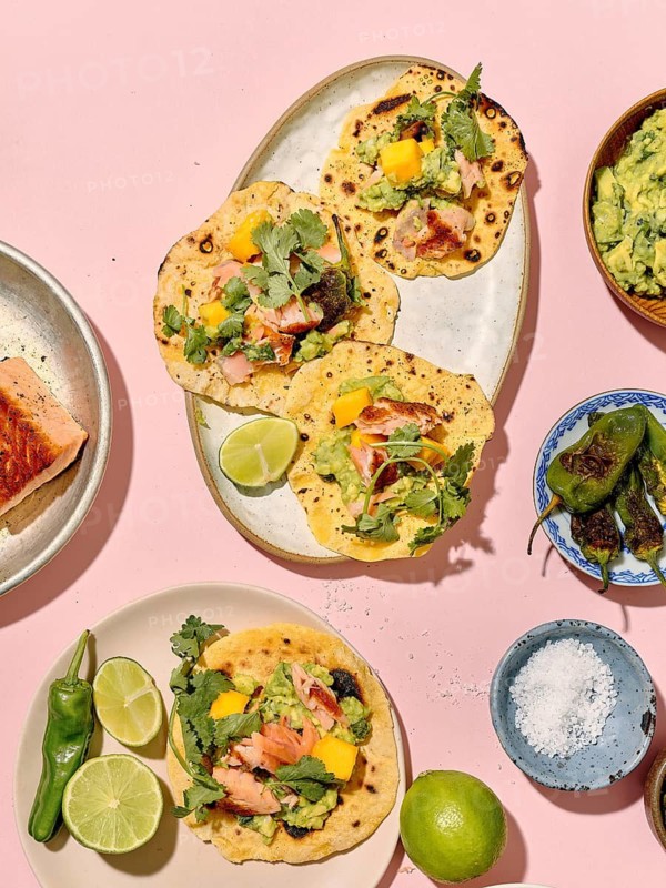 Top view of vibrant, colorful tacos arranged creatively on a pastel pink background. The ingredients include grilled salmon, fresh coriander, mango slices, avocados, and lime wedges, presenting a tempting and healthy meal option