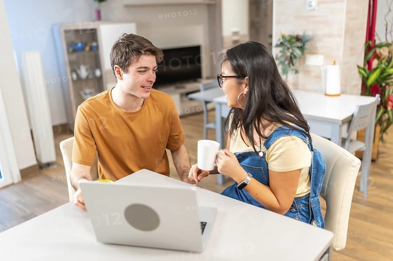Young multiethnic couple enjoying coffee and working on a laptop at their kitchen table, sharing a moment of connection in their modern home