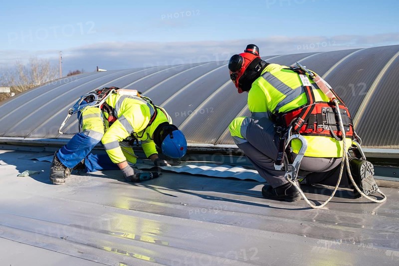 Two roofers wearing helmets and harnesses are carrying out sealing work on a flat roof near a skylight in Poland