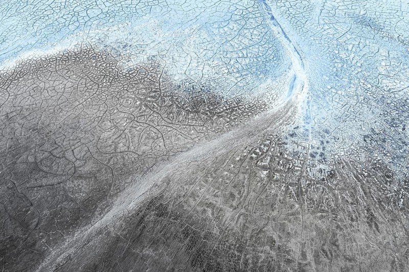 A stunning aerial view showcases intricate patterns of cracked earth and icy formations, blending shades of blue, gray, and white, creating an abstract natural landscape