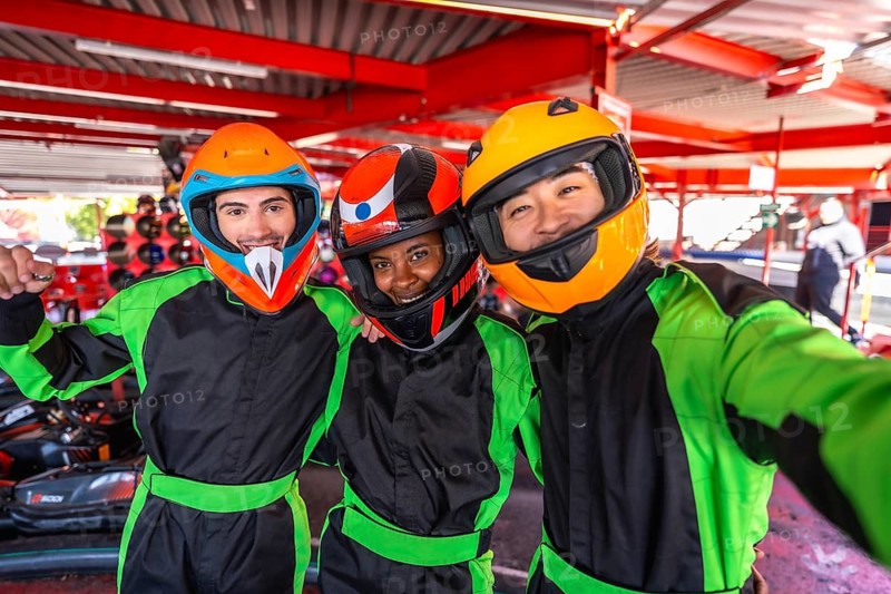 Diverse friends wearing helmets and racing suits, smiling and taking a selfie together at a go kart track, celebrating friendship and fun before a thrilling race