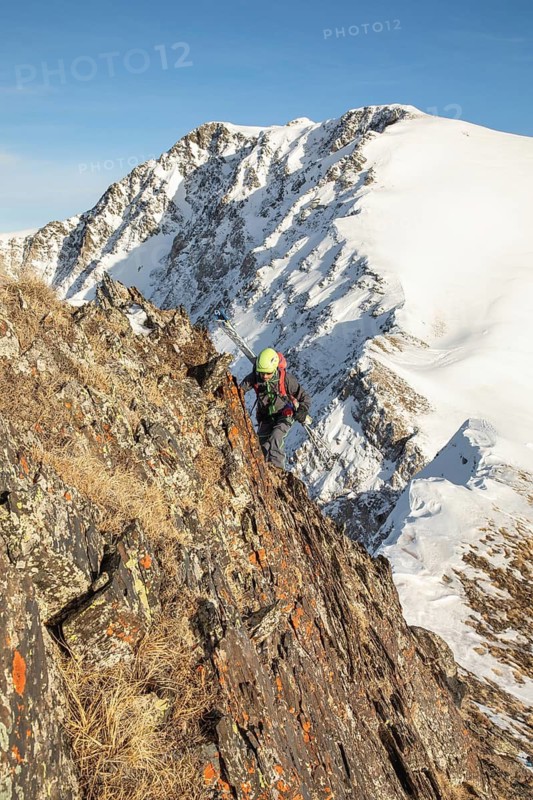 A ski tourer navigates a rocky mountain ridge, surrounded by snow-capped peaks under a clear blue sky, showcasing the thrill and challenge of alpine exploration