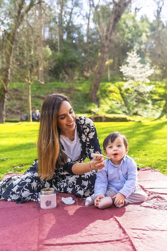 Happy mother feeding her baby solid food with a spoon while having a picnic lunch outdoors in a park