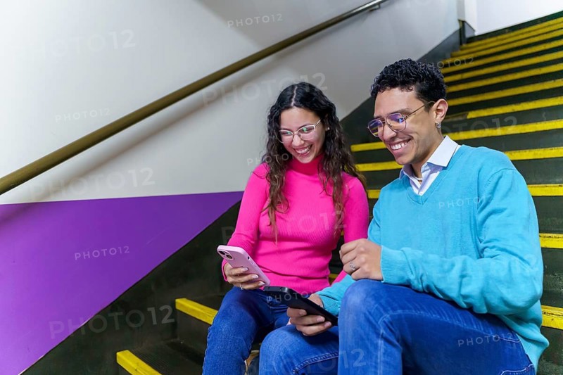 A cheerful smiling young woman and man, both Gen Z, smile while using a smartphone and tablet on a staircase, showcasing modern digital communication and connection