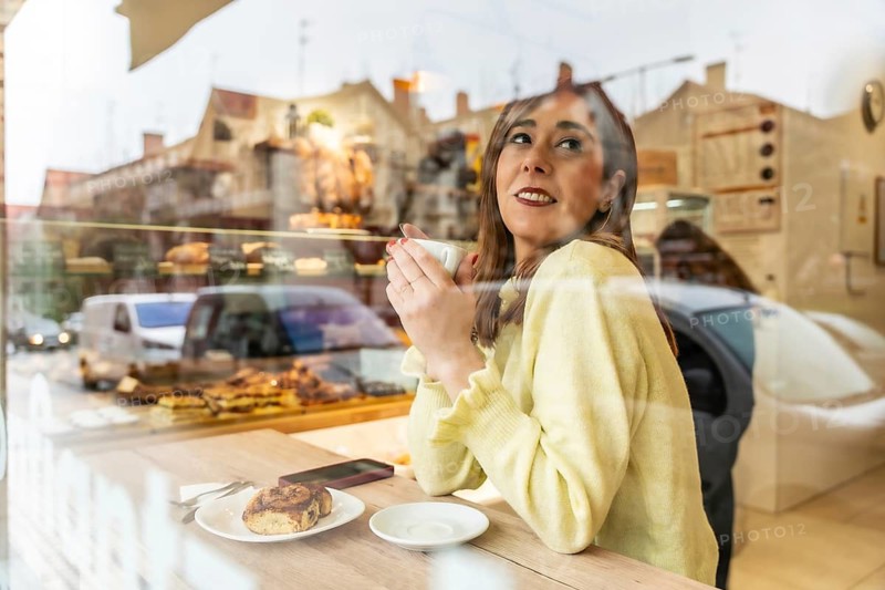 Young woman enjoying a warm coffee and sweet pastry inside a bakery, looking out through the window with reflections of city life, relaxing and contemplating