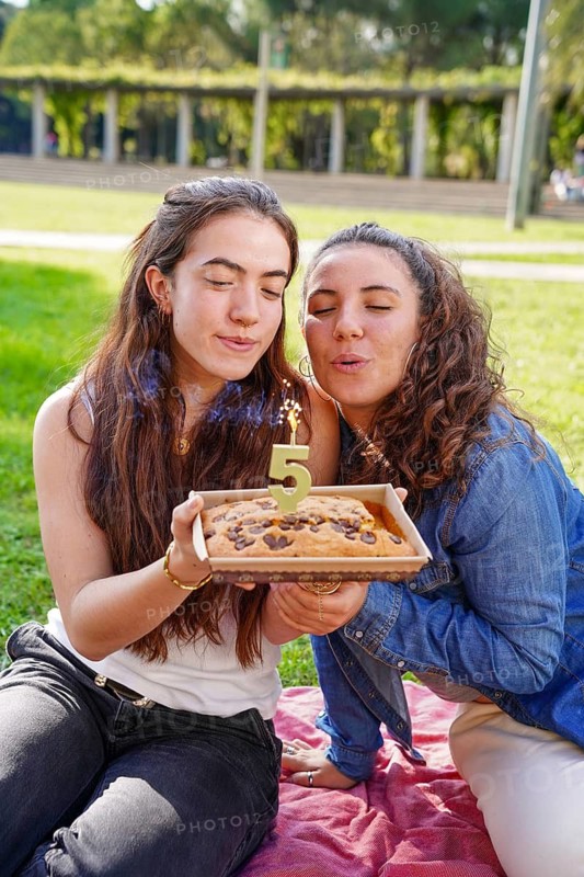 A romantic young women couple blows out a '5' candle on a cookie anniversary cake while sitting on a picnic blanket in a sunny park, exemplifying joy and love in an lgtbiq+ relationship