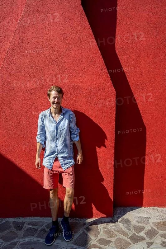Young man leaning against a red house wall, colourful houses on the island of Burano, Venice, Veneto, Italy