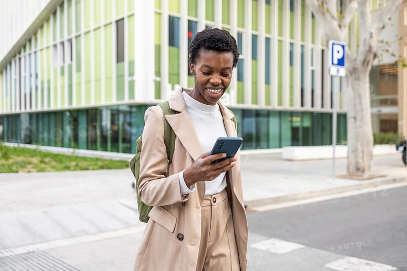 A black woman walks through an urban setting, smiling as she checks her smartphone She wears a stylish beige outfit, embodying confidence and modernity in a vibrant cityscape