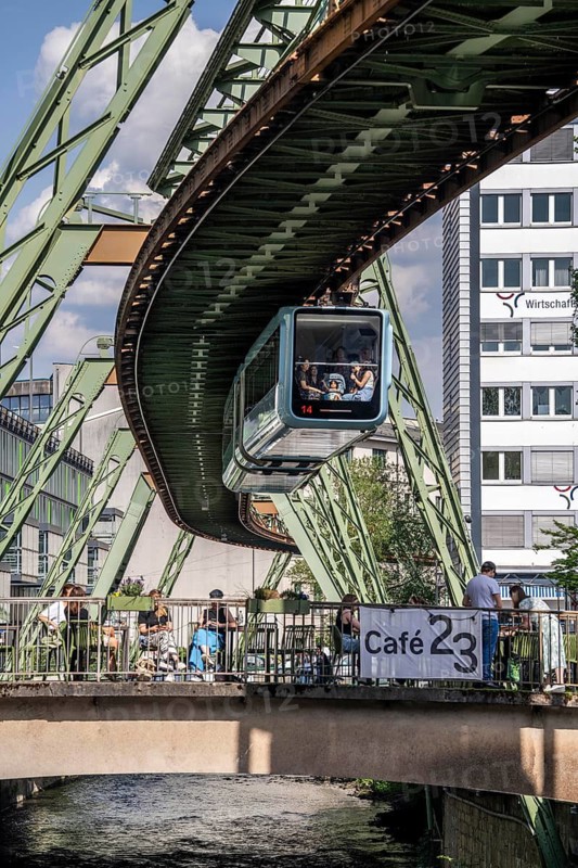 Train of the Wuppertal suspension railway runs along the Wupper, at the main station stop, terrace of Cafe23 on a small bridge over the river, North Rhine-Westphalia, Germany