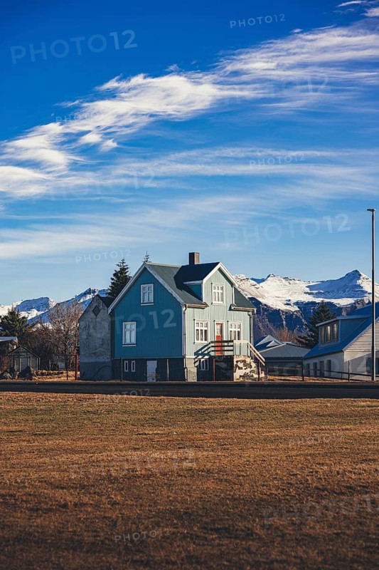 Traditional Icelandic house with a teal-blue facade and red accents stands in front of a snow-capped mountain range near Stokksnes, under a vibrant blue sky