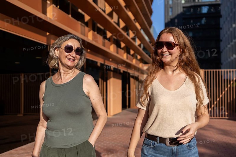 A mom and daughter enjoy a sunny day in the city, both wearing casual outfits and sunglasses. They are walking and smiling, highlighting a joyful urban lifestyle