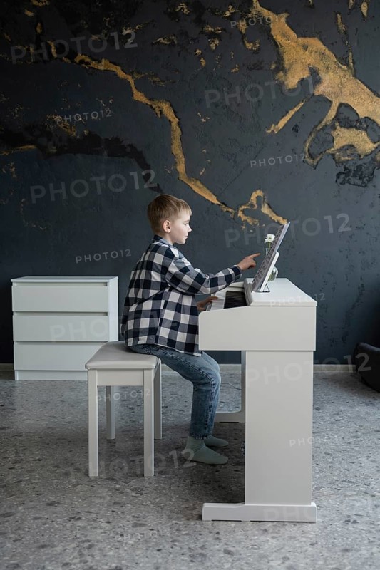A young child sits at a sleek white piano, focused on playing music. The modern interior features a stylish dark wall, creating a contrast with the bright piano