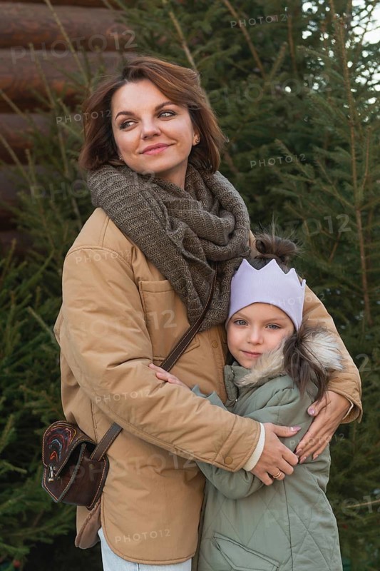 A woman and a young girl, tightly embracing outside near a Christmas tree, radiating happiness and warmth in their winter attire. The woman looks away contently while the girl looks directly at the camera, both enjoying a family celebration