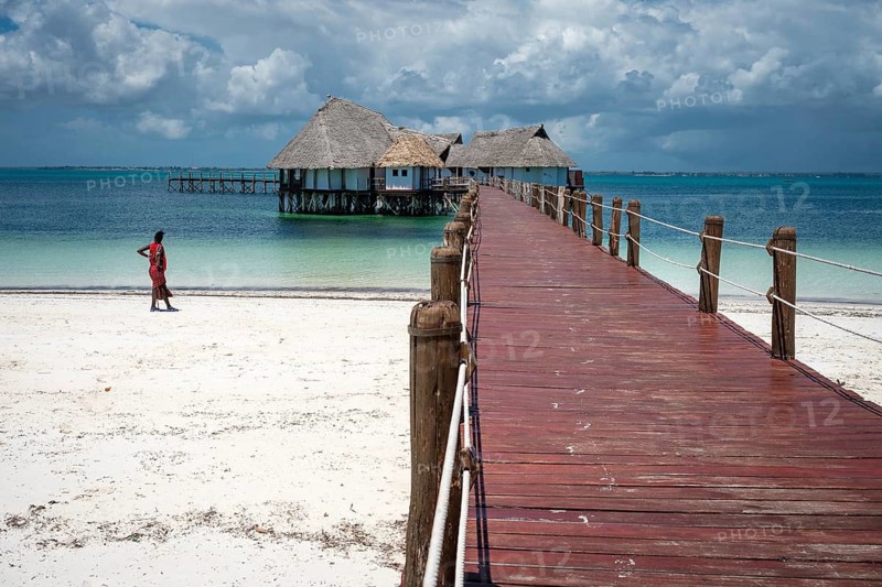 A long wooden pier leads to traditional thatched-roof bungalows over turquoise waters on a white sandy beach under a cloudy sky in Zanzibar