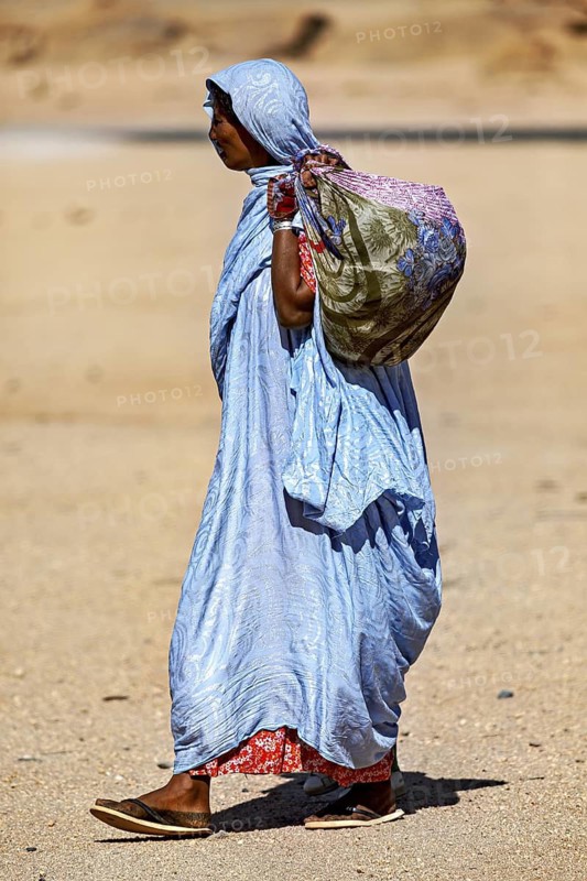 Woman in traditional dress with bag walking through a dry, sandy desert landscape, woman in the Sahara in Algeria