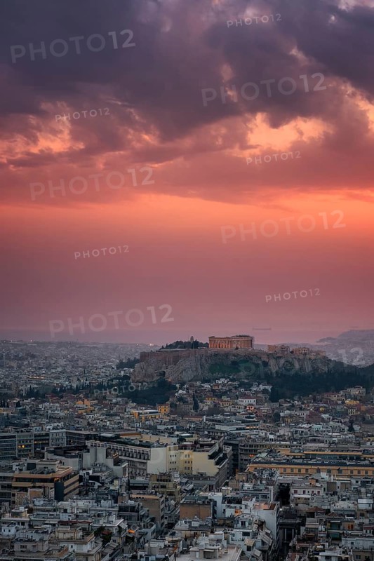 A breathtaking view of Athens at sunset, featuring the Parthenon and Acropolis illuminated against a dramatic sky. The cityscape extends towards the horizon, creating a stunning urban panorama