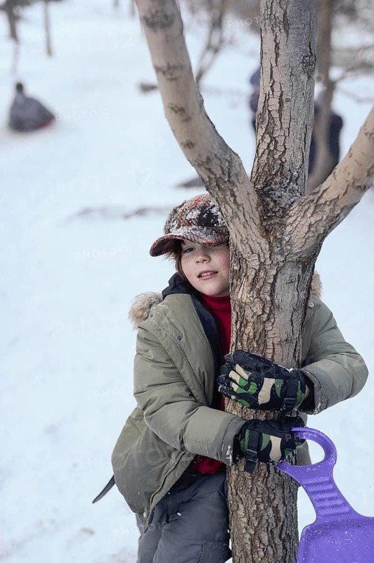 A child enjoys a snowy day at Sierra Nevada Ski Resort, Granada. Dressed warmly, they hug a tree, holding a purple sled, surrounded by a winter landscape