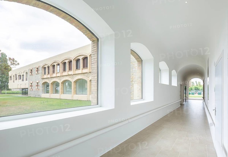A bright corridor with arched windows offers a view of a historic hospital stone facade. Natural light enhances the serene atmosphere and architectural details