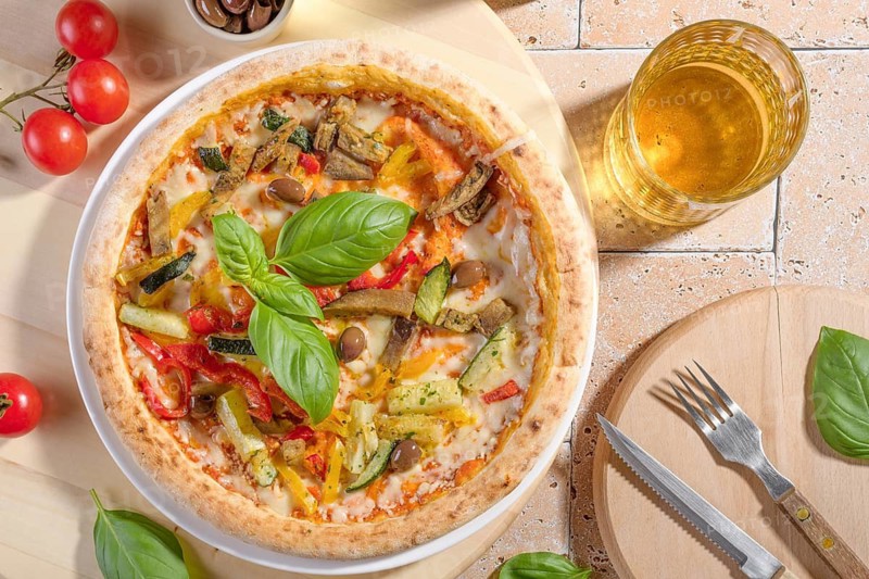 Closeup of a vegetarian pizza topped with fresh basil leaves, zucchini, eggplant, olives, and colorful peppers. Served on a rustic table with a refreshing drink on the side