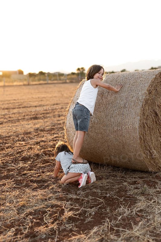 Two children enjoy a sunny day in a rural field. One climbs a large hay bale while the other crouches, experiencing playful outdoor fun and adventure in the warm evening light