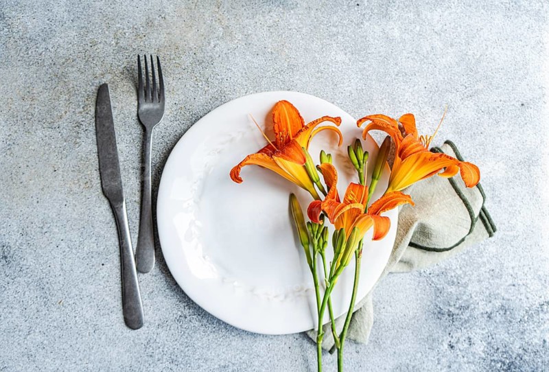 From above, a set of vibrant orange tiger lily flowers artfully adorns a white dining plate, accompanied by a knife and fork on a stone textured background
