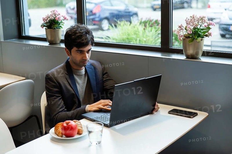A indian man works on a laptop at a hotel cafe table in a conference of AI in Amsterdam. A breakfast of croissants and apple sits beside a smartphone and a glass of water