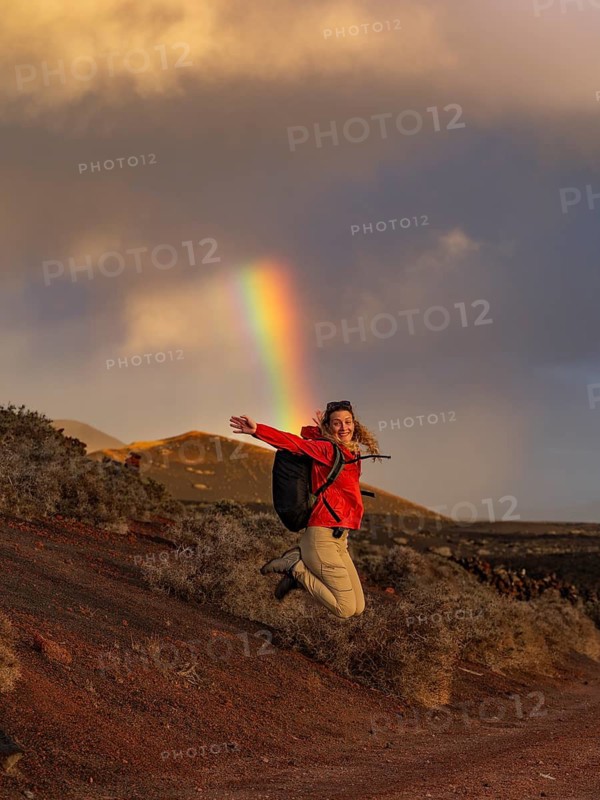 A woman in a red jacket jumps joyfully against a backdrop of a vivid rainbow in Timanfaya National Park, Lanzarote. The vibrant rainbow contrasts with the rugged volcanic landscape