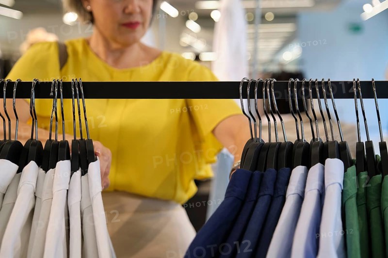 Mature woman browsing colorful shirts on hangers in a clothing store, focusing on the variety of colors and styles available, while making a thoughtful selection