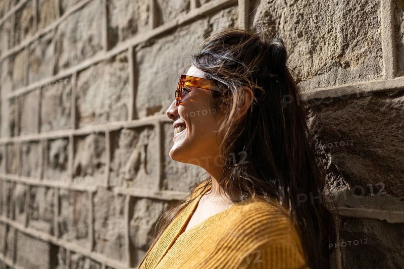 A Latin woman wearing sunglasses and a yellow sweater smiles while leaning against a textured stone wall, basking in the warmth of sunlight on a pleasant day