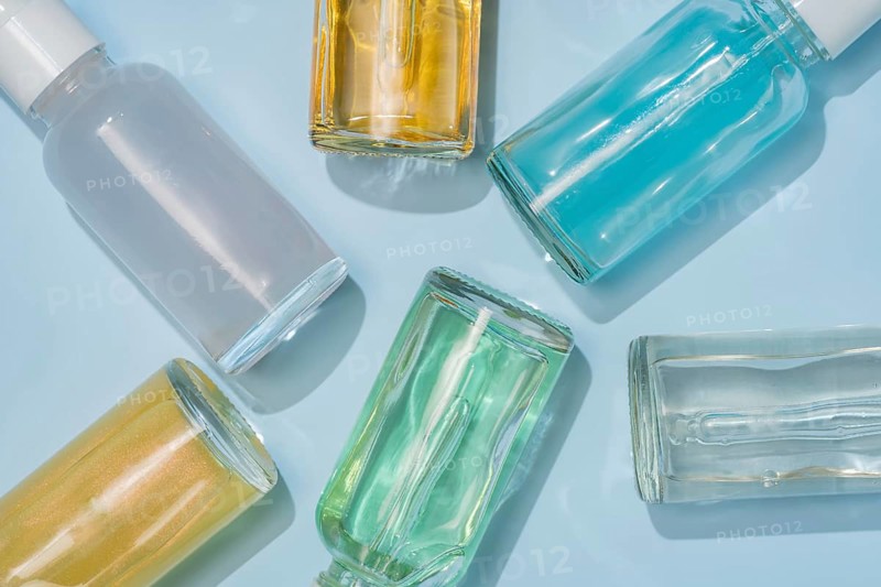 A collection of colorful glass serum bottles arranged on a light blue background. The bottles contain various vibrant liquids, showcasing a variety of hues and reflections