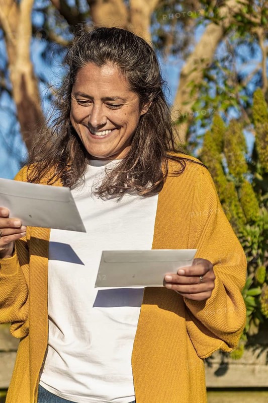 A woman stands in a sunlit Australian garden, smiling while holding two letters. Her yellow sweater contrasts with the green plants, creating a warm, inviting atmosphere