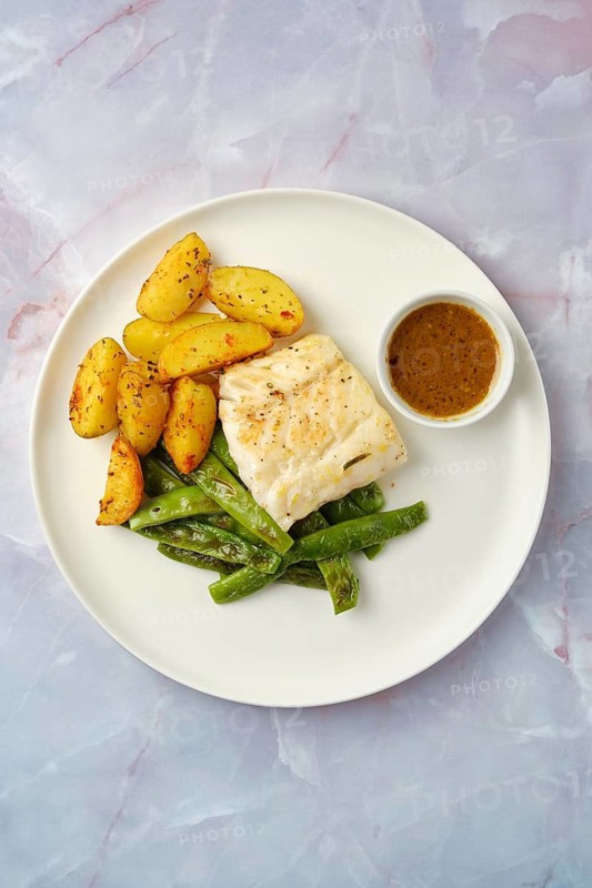 Overhead view of a plate with golden roasted potatoes and vibrant green beans accompanied by a roasted white fish fillet. A small bowl of flavorful sauce adds a touch of richness to the meal
