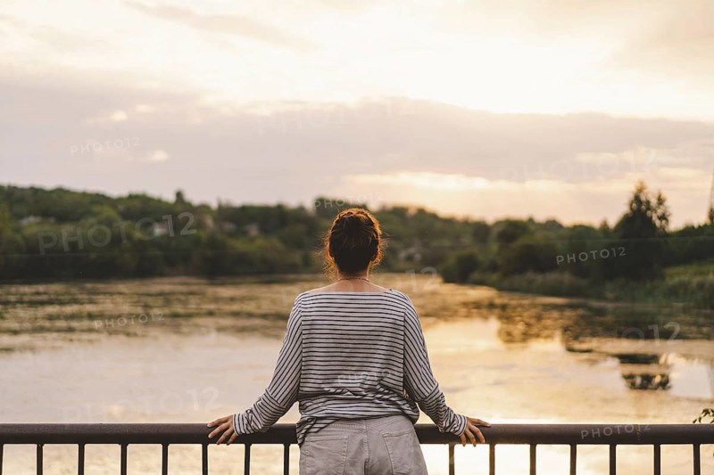 A woman stands at a railing, gazing towards a serene lake during sunset. The scene evokes a sense of tranquility and contemplation, with soft light and calm water