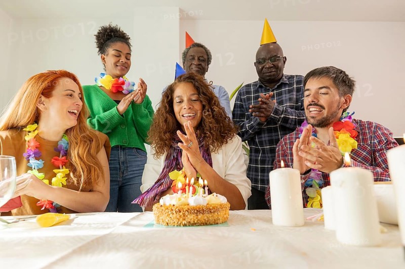 A lively birthday celebration featuring a group gathered around a cake. Party hats, colorful leis, and smiling faces create a joyful atmosphere