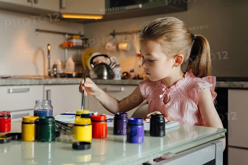 A young girl, dressed in pink, paints with bright colors at a kitchen table. Paint jars are scattered on the table, capturing a moment of creativity and focus